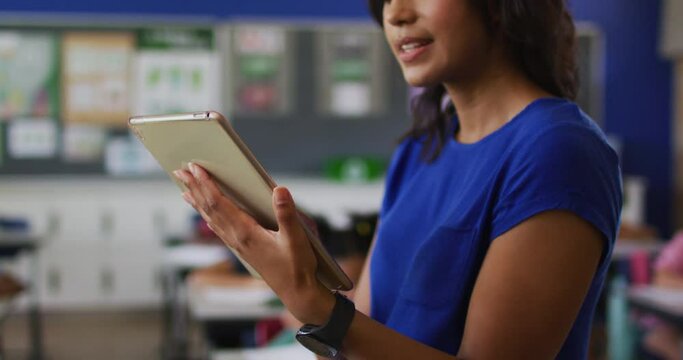 Portrait of happy mixed race female teacher standing in classroom with tablet children in background