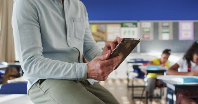 Portrait of happy caucasian male teacher in classroom with children using tablet looking at camera