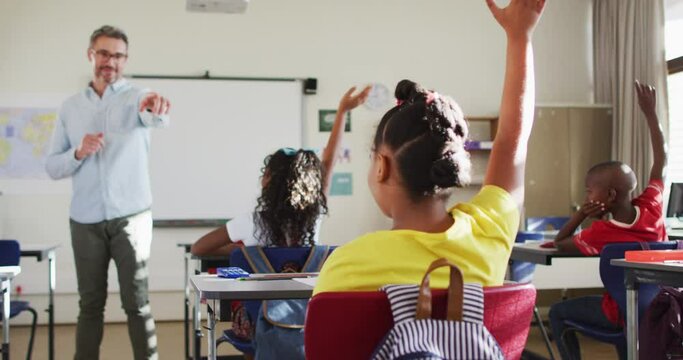 Happy caucasian male teacher in classroom with children raising hands during lesson