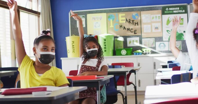 Diverse schoolchildren sitting in classroom raising hands during lesson, all wearing face masks