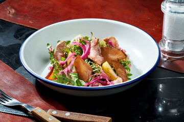 Salad with pear, onion and smoked duck in a white plate on a dark background. Hard light