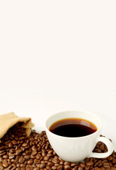 Cup of hot coffee with roasted coffee beans scattered from burlap bag on white background