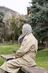 Grandma is sitting on the concrete  and looking at the hill in the background. An older woman with gray hair wears a cream coat and a scarf outside