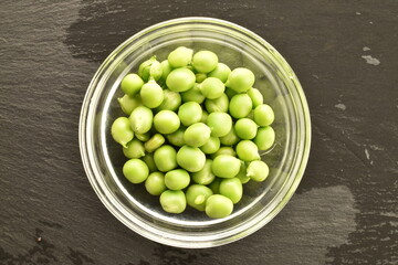 Several ripe organic, green peas in a glass dish on a slate board, close-up, top view.