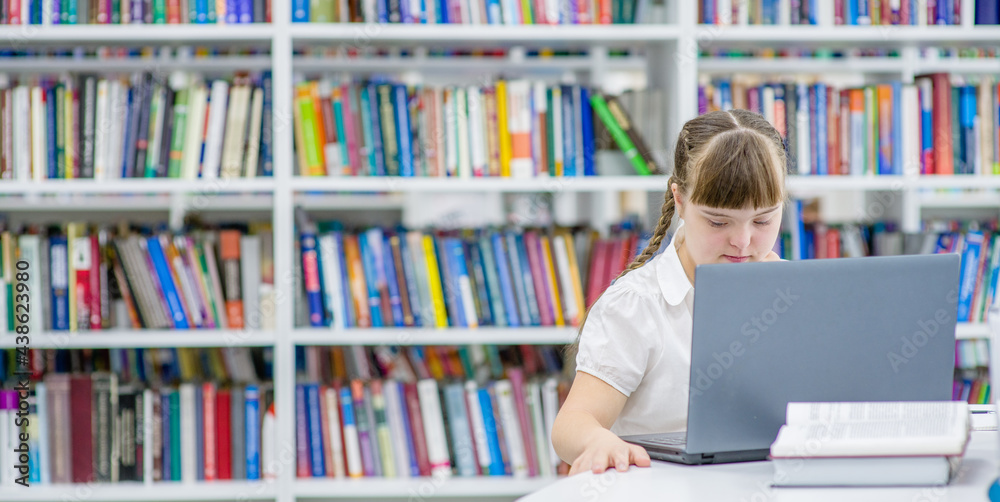 Wall mural Pensive girl with syndrome down uses a laptop at library. Education for disabled children concept. Empty space for text