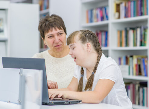 Young Girl With Down Syndrome  Uses A Laptop With Her Teacher At Library. Education For Disabled Children Concept