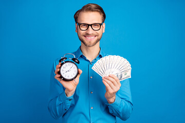 Photo of young business man happy positive smile hold clock time cash rich dollars isolated over...