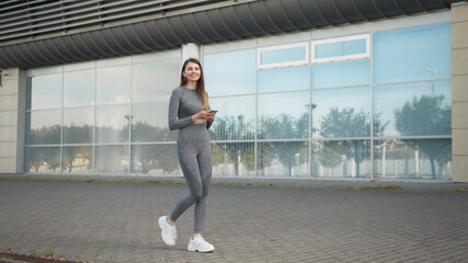 Fototapeta na wymiar Sporty beautiful young girl walks slowly in the city holding a phone in her hands and smiles. Joyful caucasian fitness trainer woman using mobile phone social media smiling while.