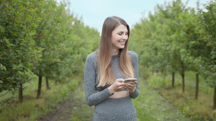 Happy woman typing by mobile phone outdoors. Cheerful girl with smartphone in park on a background. Smiling lady holding cellphone in hands outside. Athletic fitness girl holding a phone in her hands.