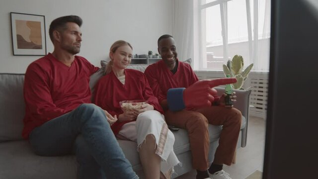Steadicam of three Caucasian and African men and woman wearing matching red long-sleeved t-shirts, sitting on couch at home, watching game, talking