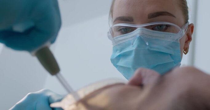 Close-up a female dentist in a disposable mask and eyeglasses doing dentist treatment to the male patient who is laying with optragate in the mouth while an assistant using a saliva ejector.