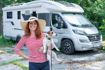 Caucasian red-haired woman in a straw hat and striped sweater walks with a small dog in front of a mobile home. Travel in a camper van with your pet