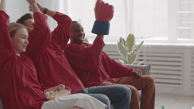 Medium long side view of three diverse sports fans wearing matching red long-sleeved jerseys, sitting on sofa at home, raising hands up, happy for favorite team winning game