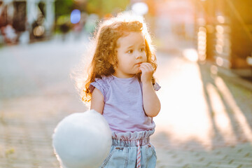 Little curly gir with white sweet cotton candy in the park at summer sunset.