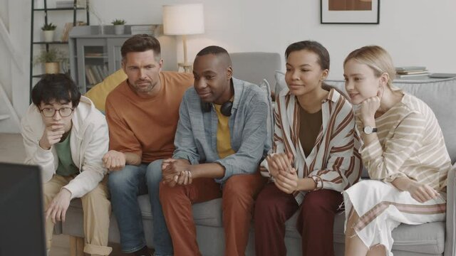 Medium long of five young multiethnic friends sitting on couch in living room at home, watching TV, Mixed-Race woman secretly tapping on shoulder of Caucasian man