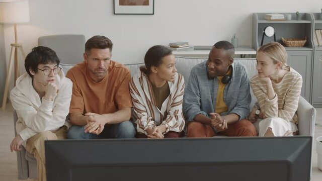 High-angle of African, Asian, Mixed-Race and Caucasian women and men sitting on sofa in living room, watching television and talking