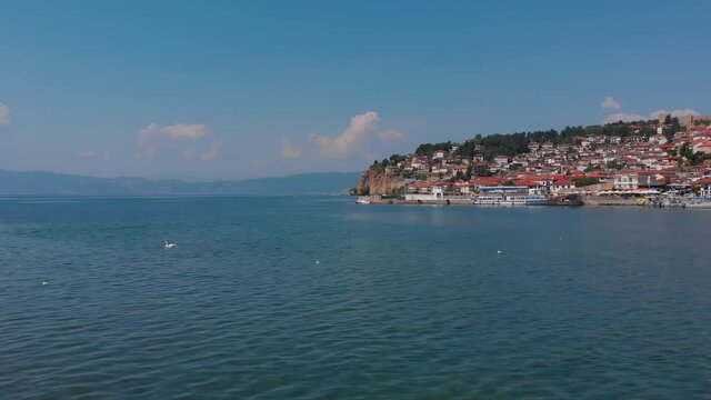 A huge lake of Ohrid, North Macedonia. Historic Medieval houses with red roofs are on the shore of blue water. Birds are swimming on surface. Beautiful scenic took with aerial drone 4K 30fps.