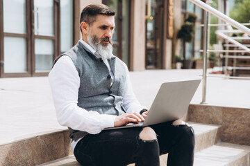 Senior man stylish man with gray beard spends time outdoors in laptop.