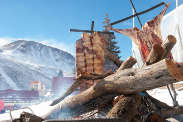 Costillar de ternero y chivo estacados en cruz para cocinar a la llama al aire libre con nieve con fondo de montaña nevada y parte de pista de sky