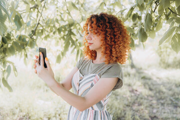 Redhead girl with afro curls with a phone in a green park makes a selfie and smiles, makes a...