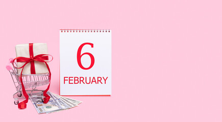 A gift box in a shopping trolley, dollars and a calendar with the date of 6 february on a pink background.