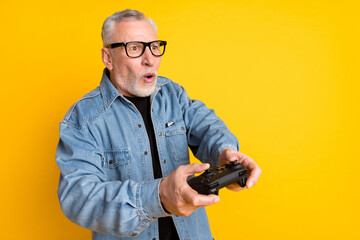 Photo of impressed feel young grandpa play video game wear eyewear jeans jacket isolated on yellow color background