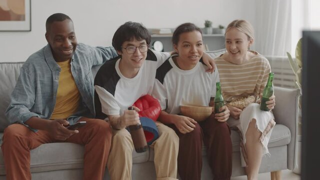 Medium long of four diverse happy young people sitting on couch at home with beer and snacks, smiling and cheering