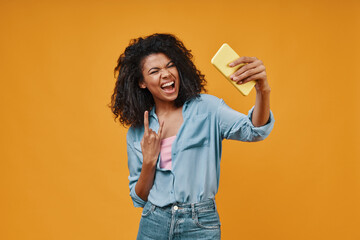 Playful young African woman in casual clothing taking selfie and gesturing while standing against yellow background