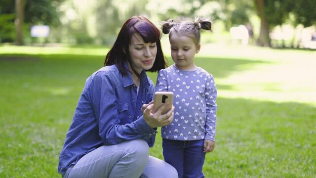 Mother and cute little 3 years old daughter, wearing casual blue clothes, taking selfie photo with phone camera, making video call to friend and waving, outdoor in the summer park on sunny day