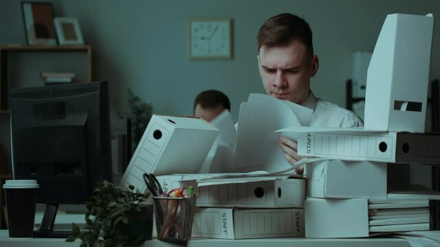 Tired irritated man with pile of folders working in office, employee having headache, exhausted worker with red eyes, pain in head, throwing documents away. Bad mood, fatigue, lots of work. 
