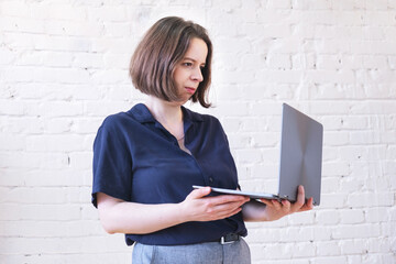 Woman in blue shirt and gray trousers stands with laptop in her hands and looks at screen. Portrait of young brunette woman. Manager is preparing for a presentation, business meeting