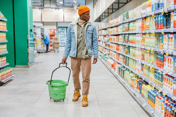 African American man in a stylish orange hat walks with an empty green shopping cart through a...