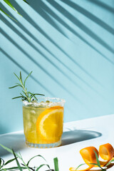 Iced orange refreshment juice with a slice inside the glass, rosemary as a garnish and a lot of ice, on a cyan background with palm tree shadows and space for text