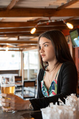 young woman with long straight hair, sitting at the table in a restaurant with wooden furniture, while holding a drink in a crystal glass, wearing casual clothes, lifestyle