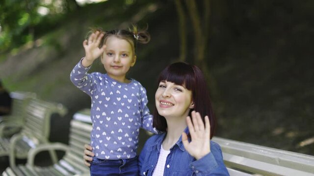 Beautiful mother and her adorable little 3-years old daughter, spending leisure time in beautiful city park, sitting on the bench and waving to camera. Summer family time, sunny days concept