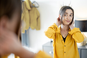 Young woman looks on the mirror and makes hairstyle, getting ready to go out at the hallway at home