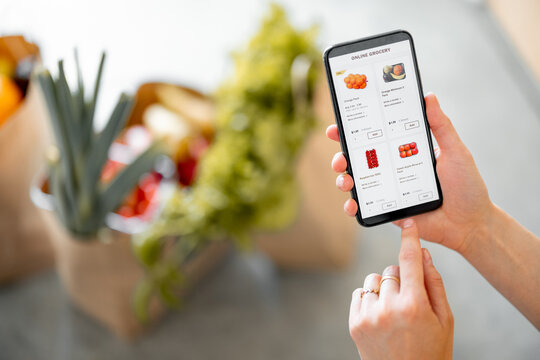 Holding phone with running e-shop application and bags full of fresh food on the background. Shopping groceries online concept