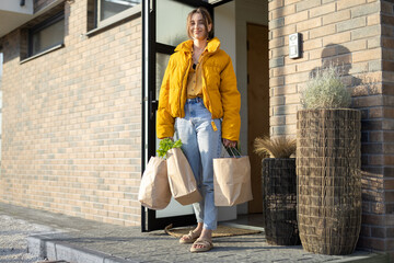 Fototapeta na wymiar Young woman standing with paper bags full of fresh groceries on the porch of her house. Shopping healthy food in eco-packaging
