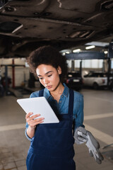 young african american mechanic standing underneath car and holding digital tablet in garage