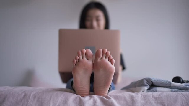 Feet of Asian woman sitting on bed with blurred smiling millennial waving and talking at video chat on laptop. Unrecognizable young manager conferencing indoors in sleeping room