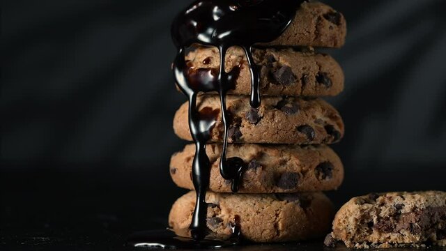 Pouring extra dark melted liquid chocolate sauce over stack of homemade cookies in slow motion. Sweet biscuits, homemade pastry, liquid texture on black background