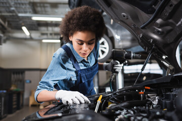 young african american mechanic in overalls holding electric screwdriver and working with car motor...