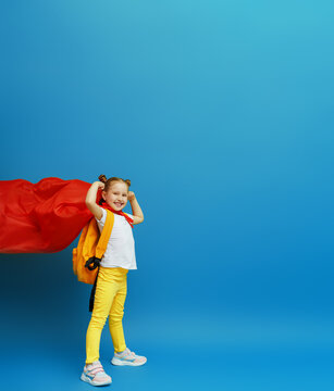 happy little schoolgirl in superhero costume with a red cape and a backpack shows off her muscles. confident, strong and successful little girl smiles and poses on a blue background. desire to study.