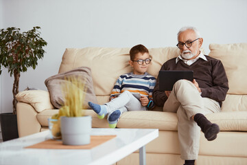 Senior man and his grandson surfing the net on touchpad while relaxing in the sofa.