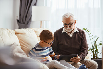 Small boy using digital tablet with his grandfather at home.