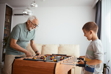 Cheerful senior man playing table foosball with grandson at home.