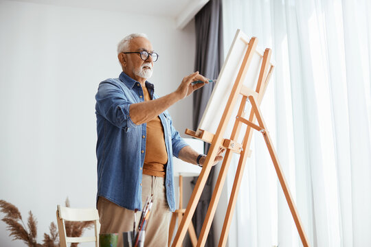 Elderly male artist painting on canvas at home.