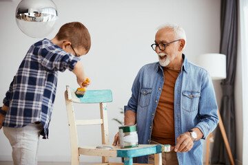 Happy grandfather and grandson having fun and painting wooden chair in blue at home.