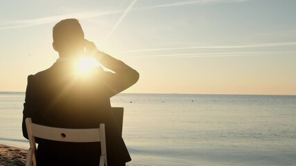 A businessman sits on a chair by the sea and works at a laptop, drinks a glass of coffee and throws an empty glass on the sand. The finish line on the way to a seaside vacation. - Powered by Adobe