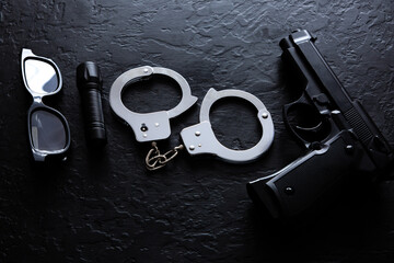 Black gun, flashlight, glasses and Police metal real handcuffs lie on the black background. Private...